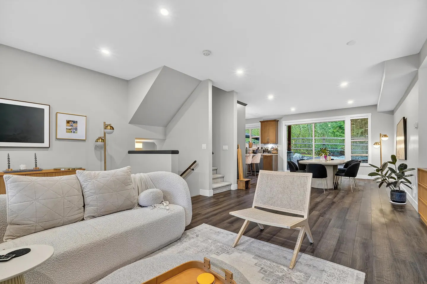 Bright, open-concept living space with a white sofa, woven chair, and dark wood floors. A dining area and kitchen are visible in the background.