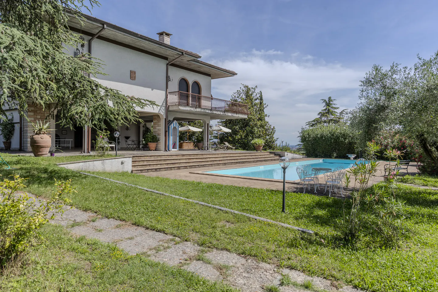 A two-story villa with a balcony, stone patio, and swimming pool set in a lush garden with trees under a blue sky.