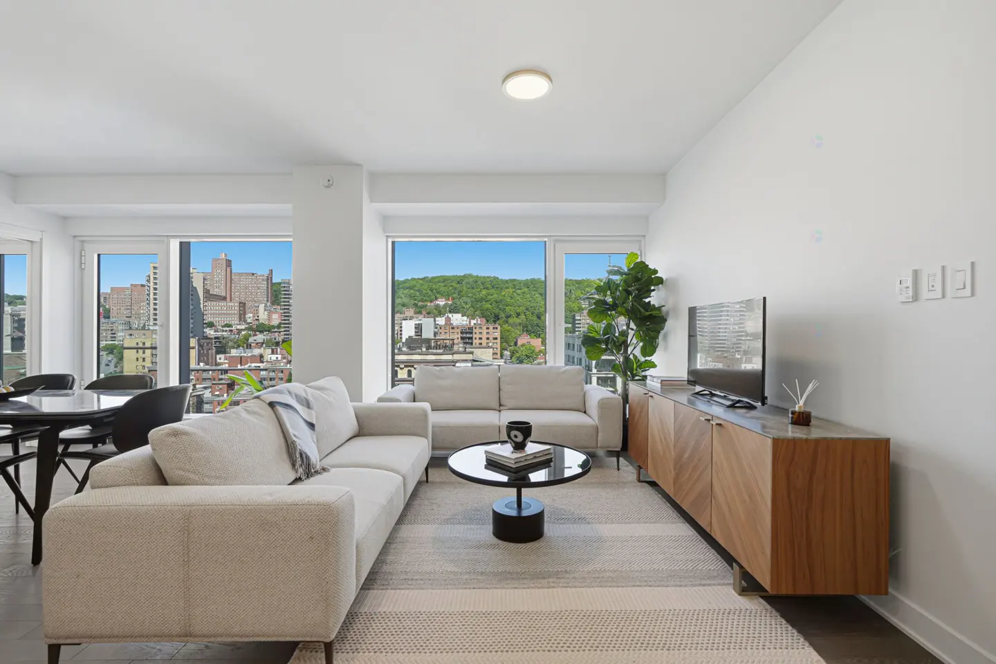 Modern living room with white sofas, a round coffee table, wood media console, and large windows showing a city view.