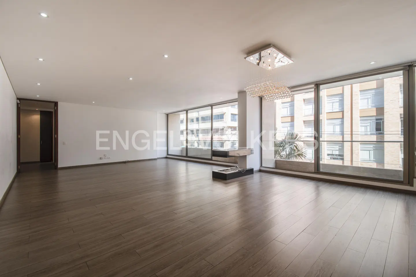 Bright, empty living room with hardwood floors, white walls, and large windows overlooking a city. A modern chandelier hangs from the ceiling.