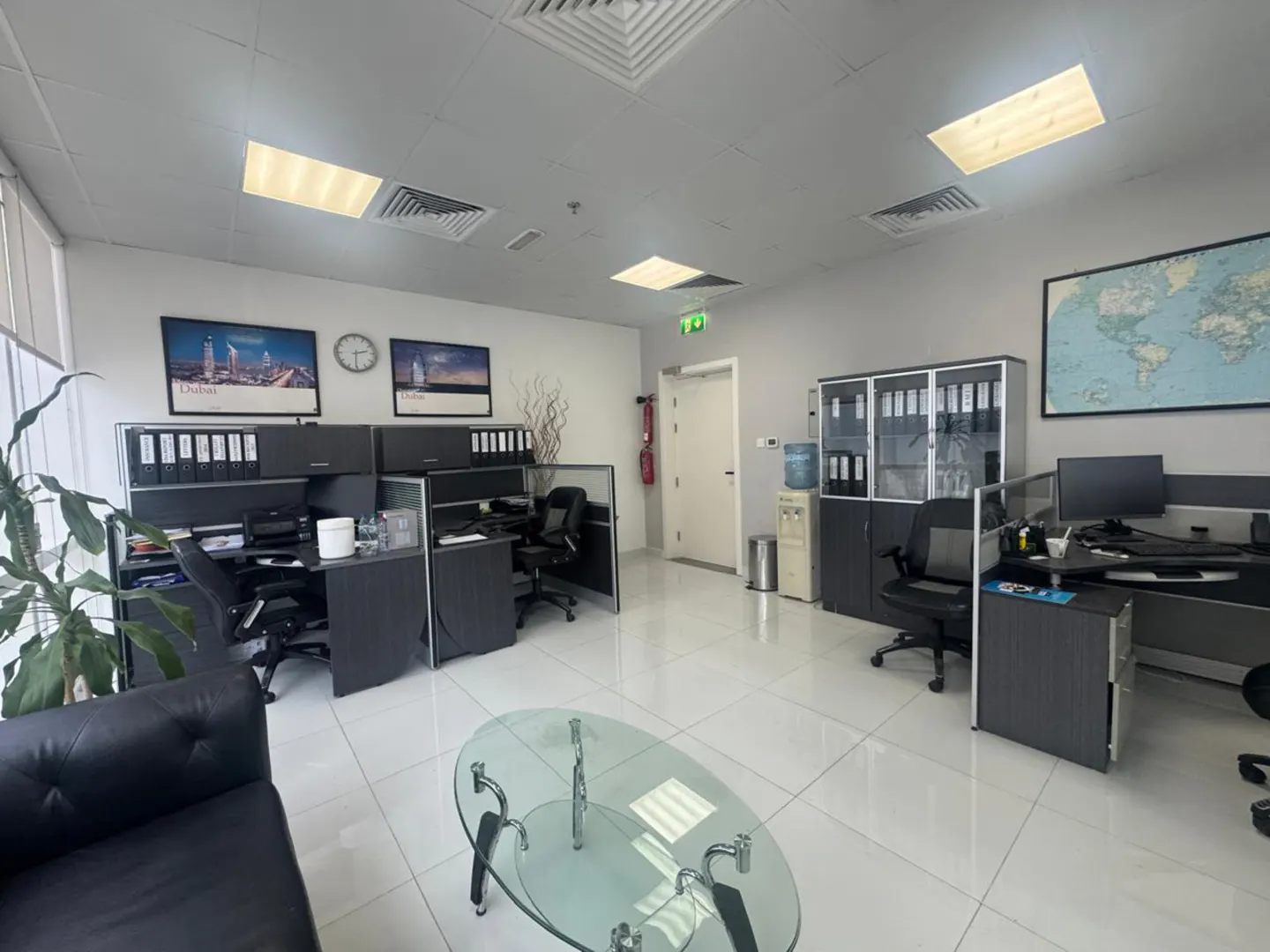 Bright office space with gray cubicles, black chairs, and white tile floors. A glass coffee table sits in front of a black leather couch.