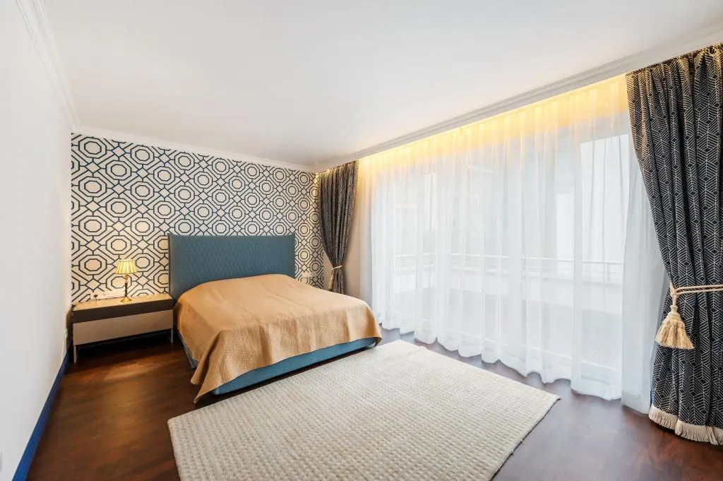 Bedroom with blue headboard, tan blanket, geometric wallpaper, and white rug on dark wood floor. Sheer curtains filter light.