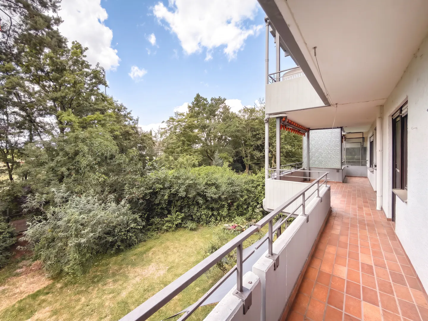 Spacious apartment balcony with terracotta tiles, metal railings, and a view of a lush green garden and blue sky.