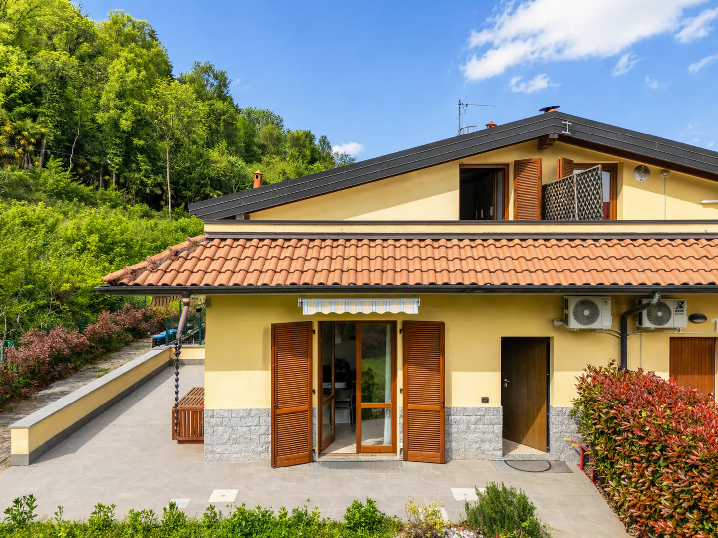 Exterior view of a yellow house with a red tile roof, brown shutters, and a gray stone foundation, set against a backdrop of green trees and a blue sky.
