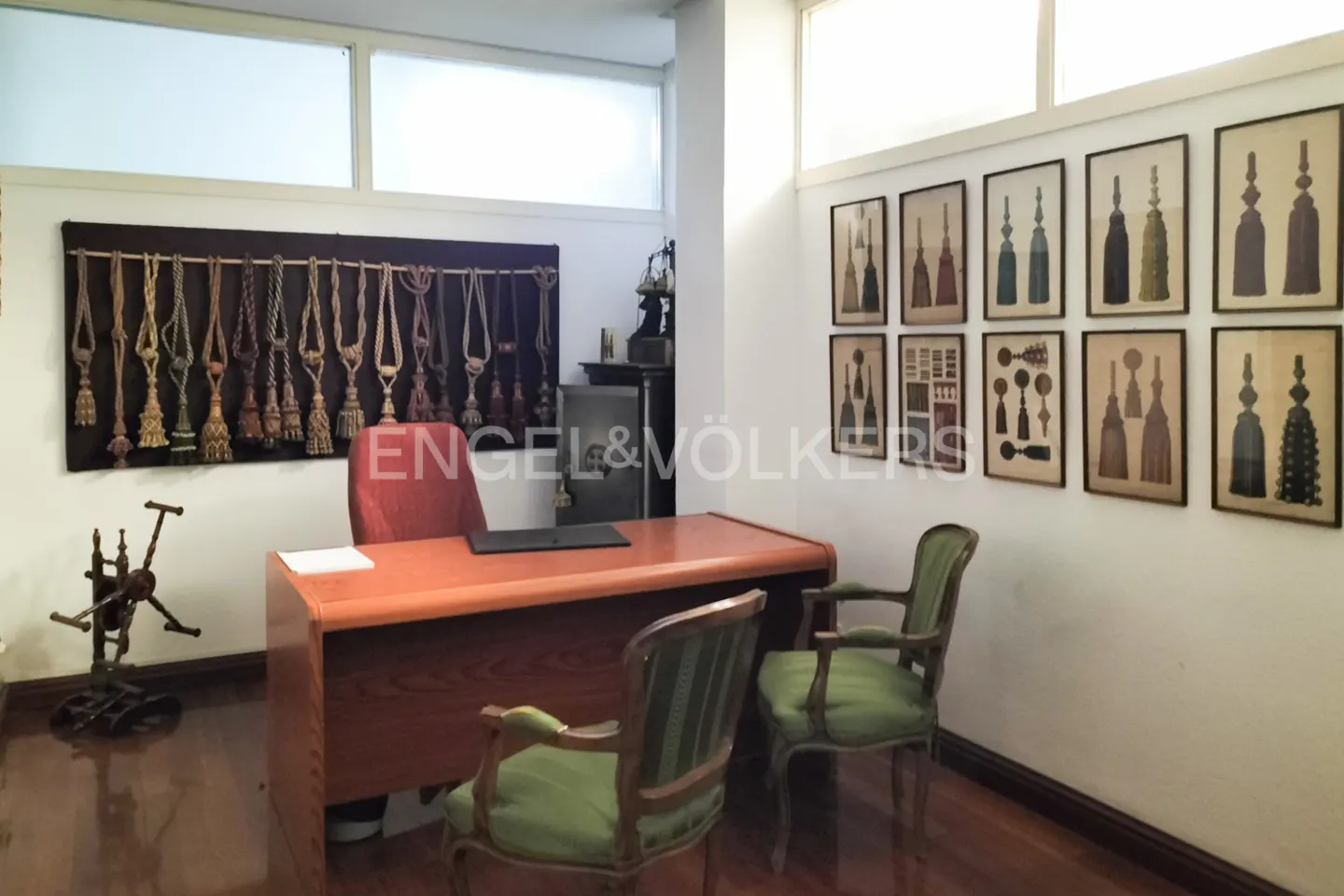 Office with a wooden desk, two green chairs, and tassel art on the walls. A spinning wheel sits in the corner.