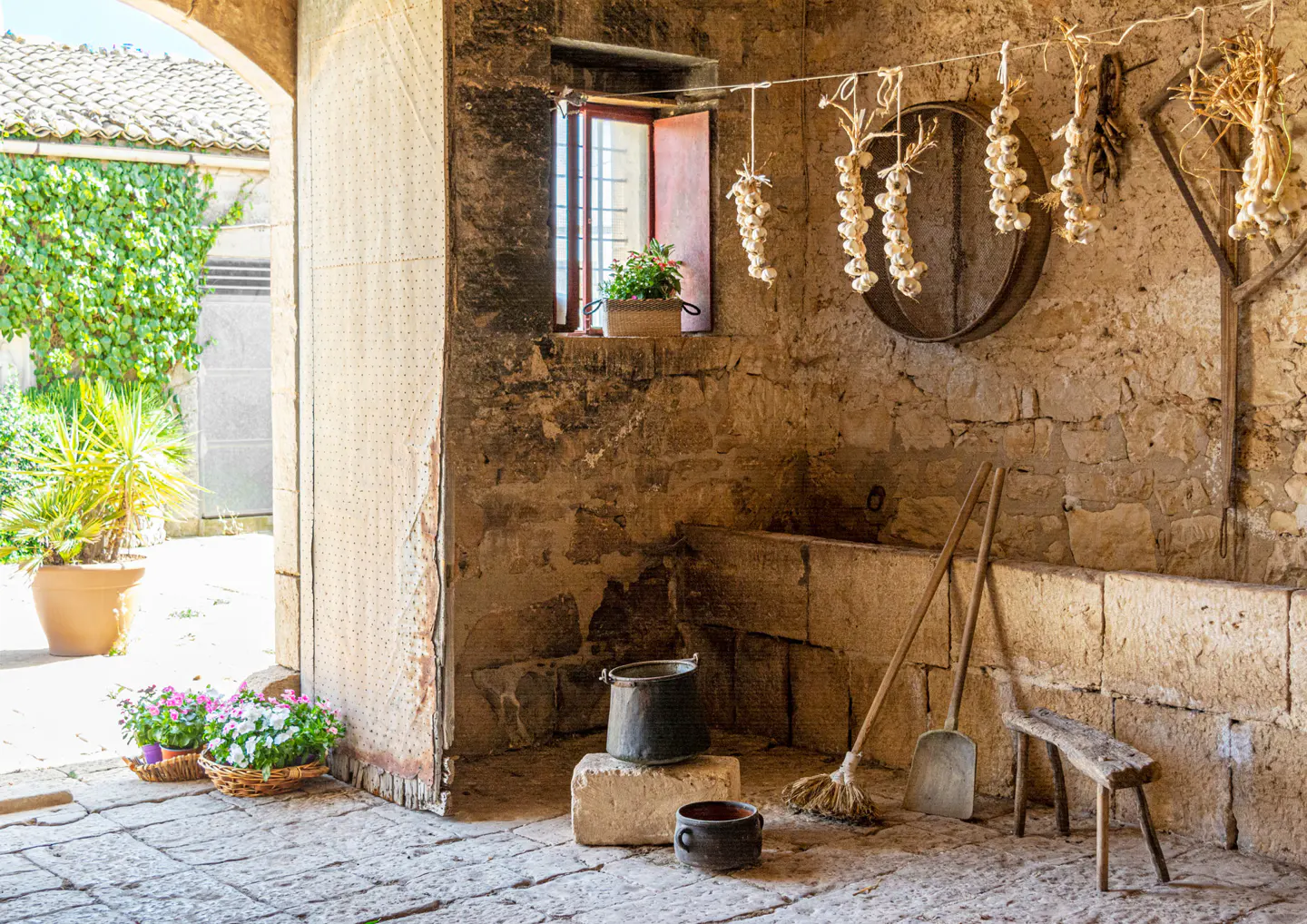 Rustic stone room with garlic hanging, tools, and flowers. An open window has a flower pot. A courtyard is visible through an archway.