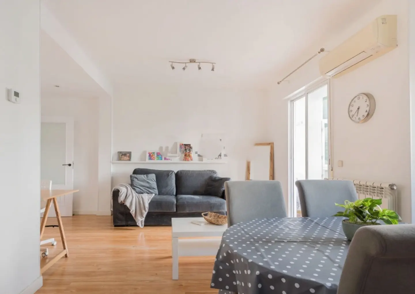 Bright living room with hardwood floors, a gray sofa, and a round table with a polka dot tablecloth. A white shelf displays photos and art.
