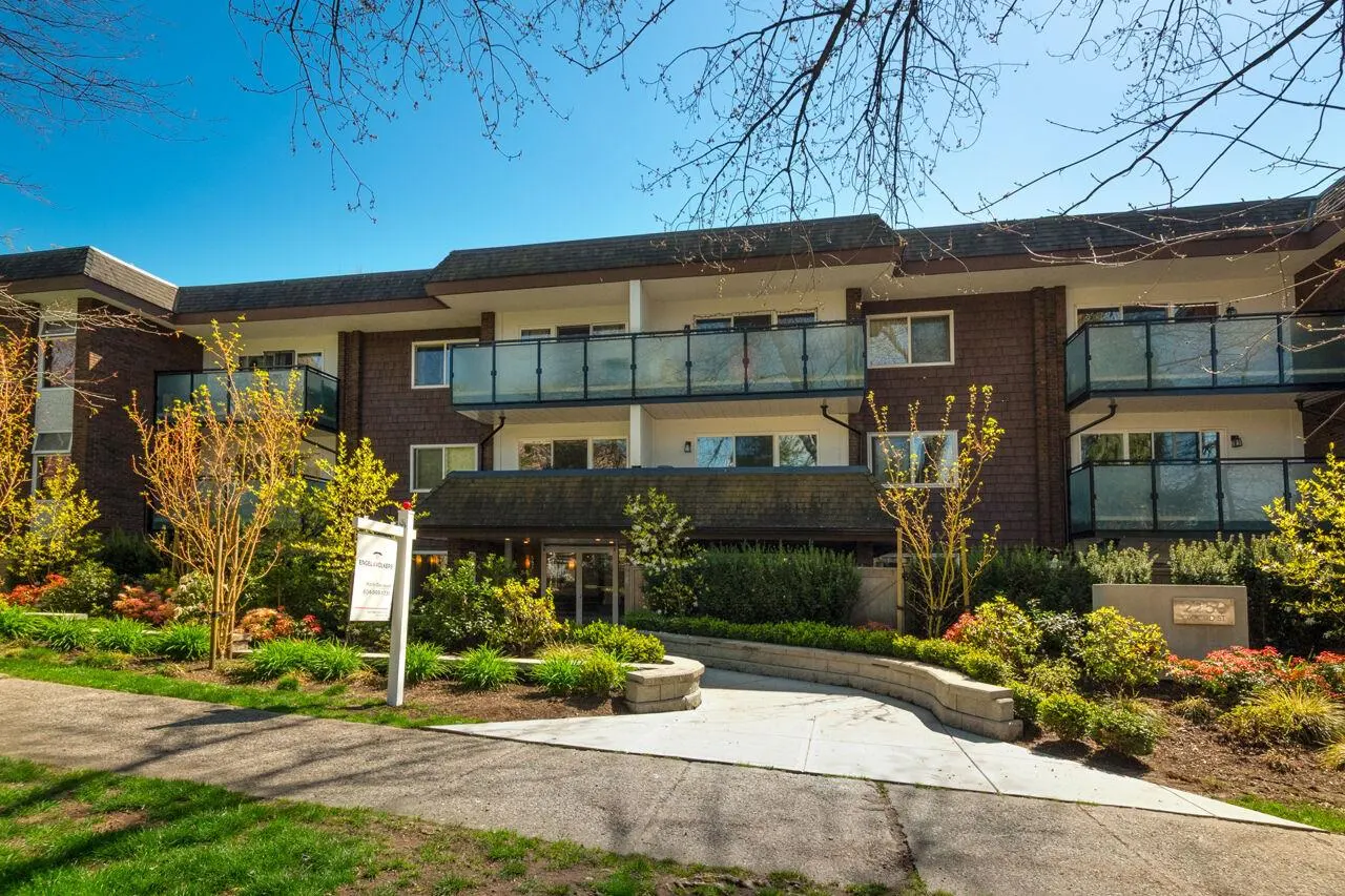 Exterior view of a two-story brown apartment building with glass balconies and a landscaped entrance on a sunny day.
