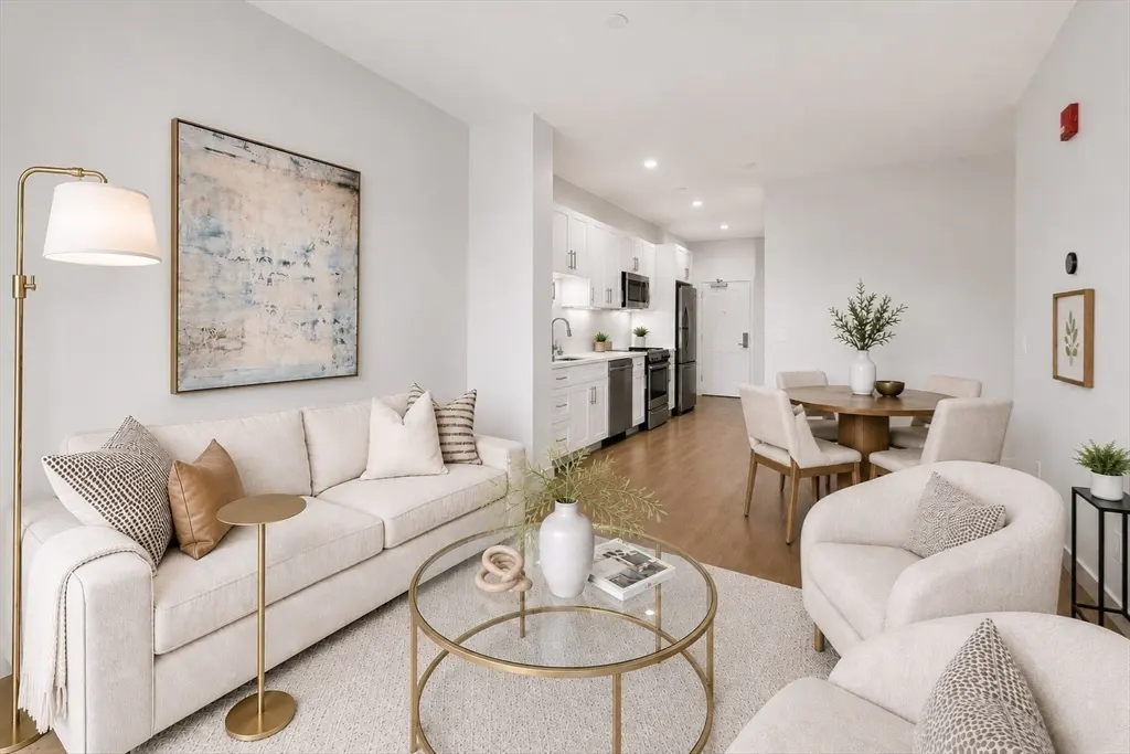 Bright, open-concept apartment with white sofa, chairs, and kitchen. A gold floor lamp and round glass coffee table add warmth.