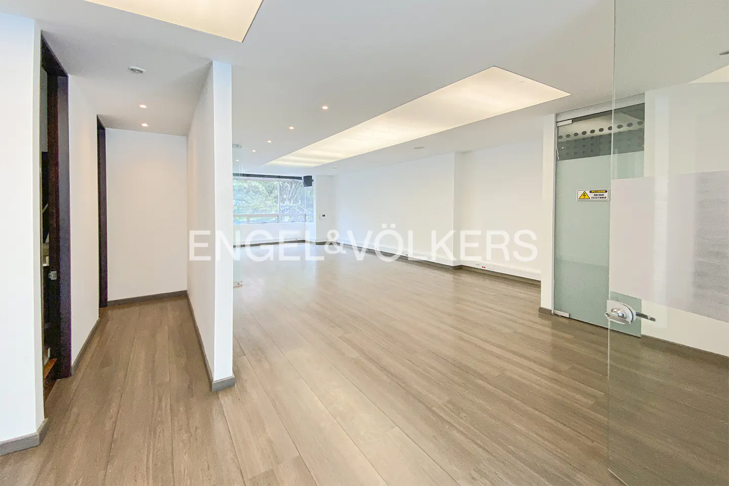 Bright, empty room with light wood floors, white walls, and recessed lighting. A glass door is on the right. Engel & Völkers logo is visible.