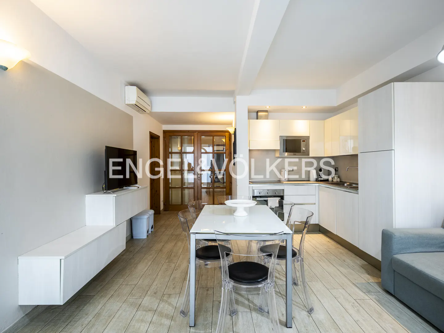 A bright, modern apartment interior with a white kitchen, dining table with clear chairs, and a TV on a white console.