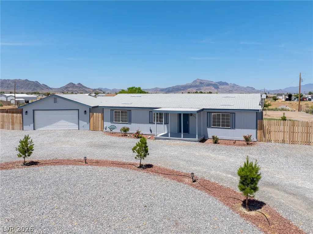 Single-story light gray house with a white garage, gravel driveway, and desert mountain backdrop under a clear blue sky.