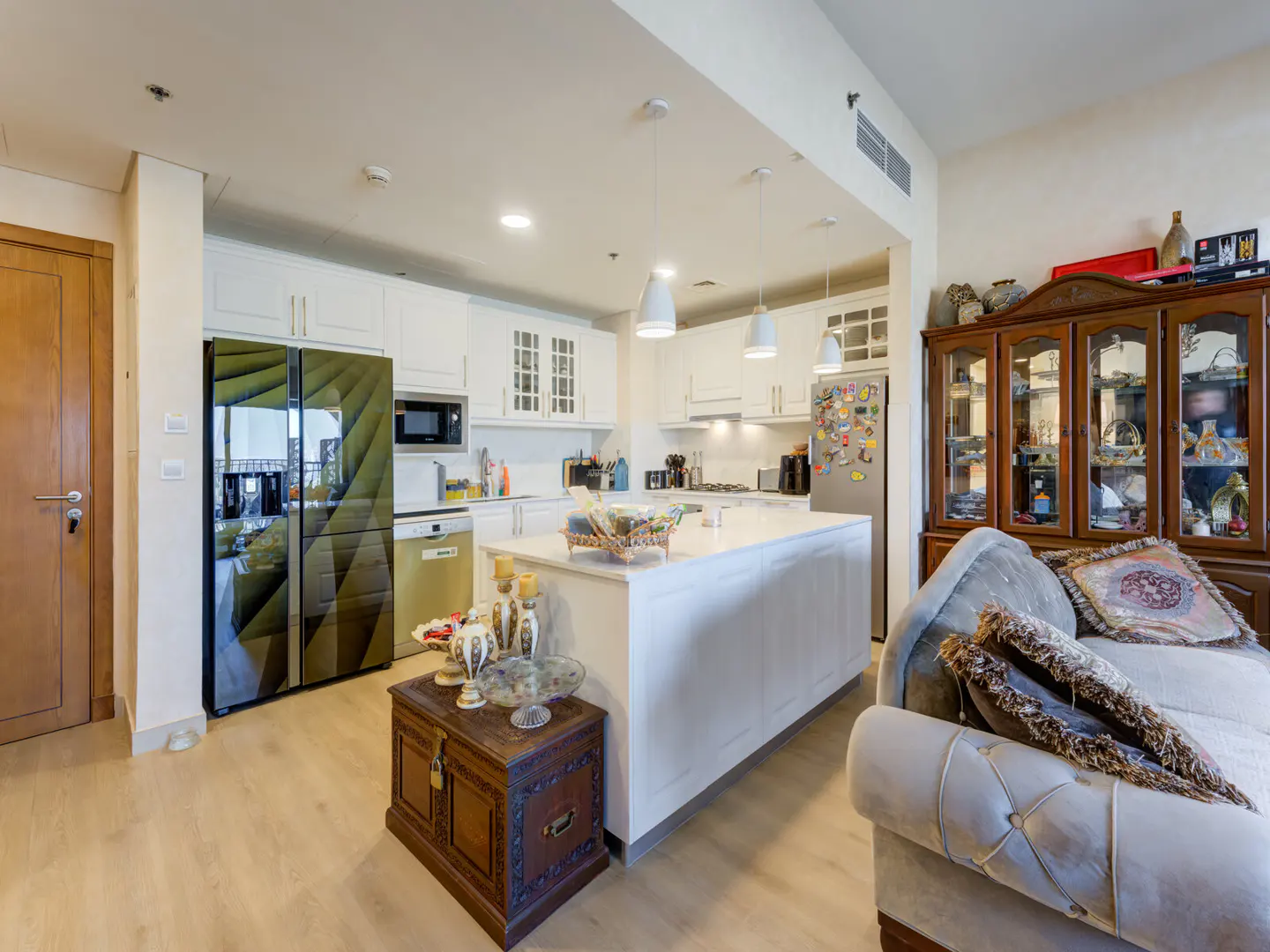 A bright, modern kitchen with white cabinets, a large island, and a unique patterned refrigerator. A wooden cabinet and sofa are in the foreground.