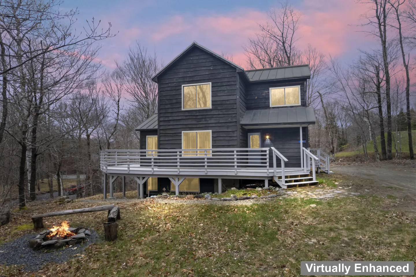 Two-story black house with a white deck, surrounded by trees. A fire pit burns in the front yard under a pink and blue sky.