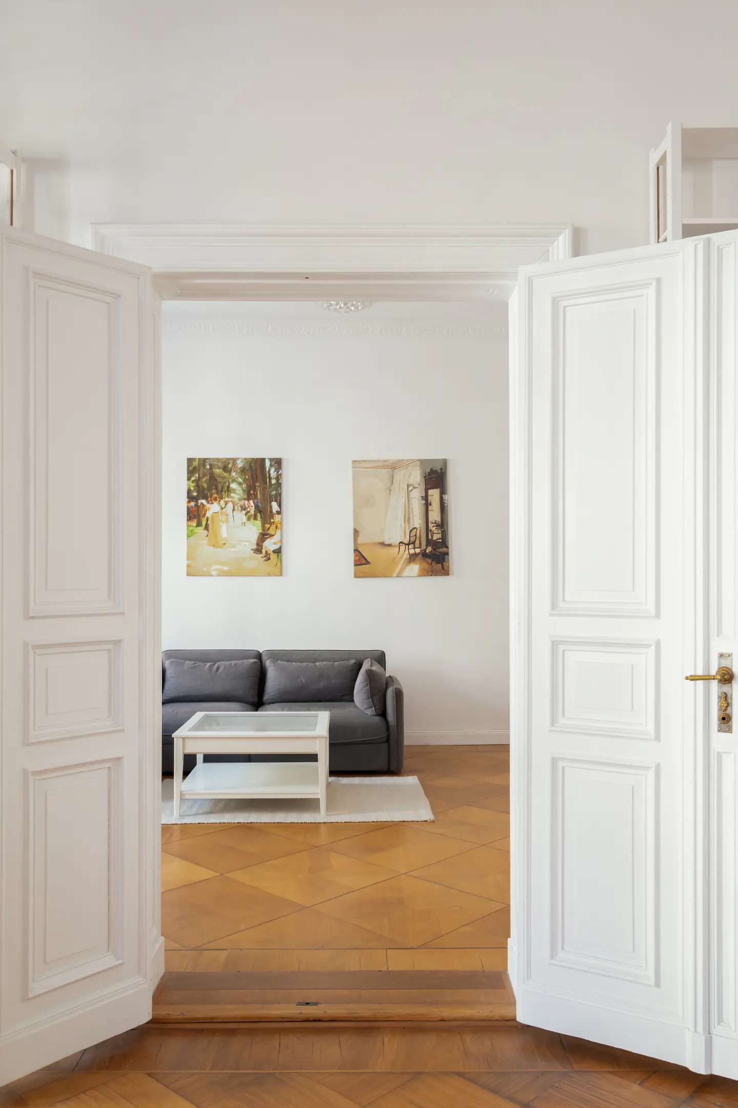 View through open white doors into a living room with a gray sofa, white coffee table, and two paintings on the white wall.