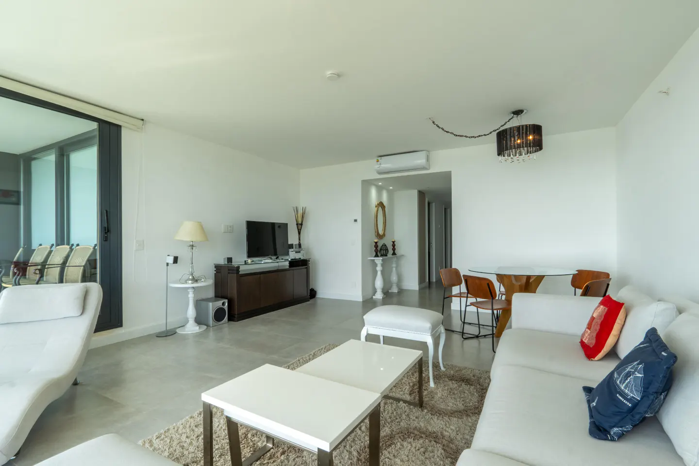 Modern living room with white sofa, two white coffee tables, dining area, and large glass doors leading to a balcony.