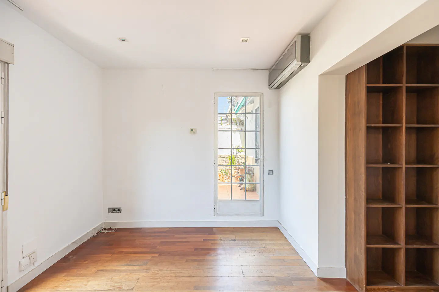 Bright, empty room with hardwood floors, white walls, and a white-framed glass door to a patio. A built-in wooden bookcase is on the right.