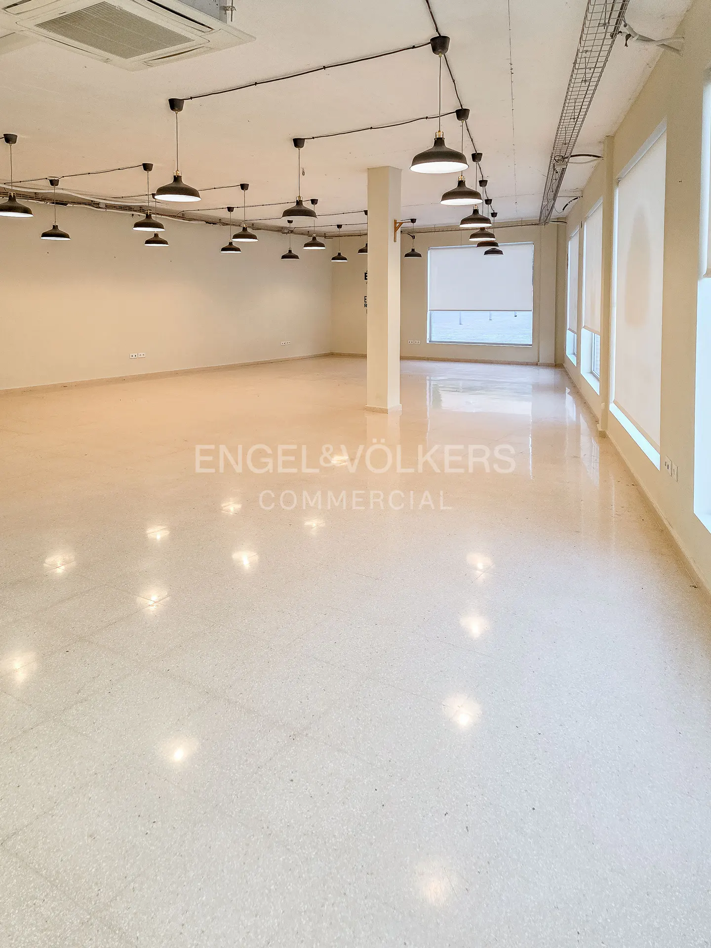 Bright, empty commercial space with white walls, speckled floor, and black pendant lights. Windows with white blinds line the right wall.