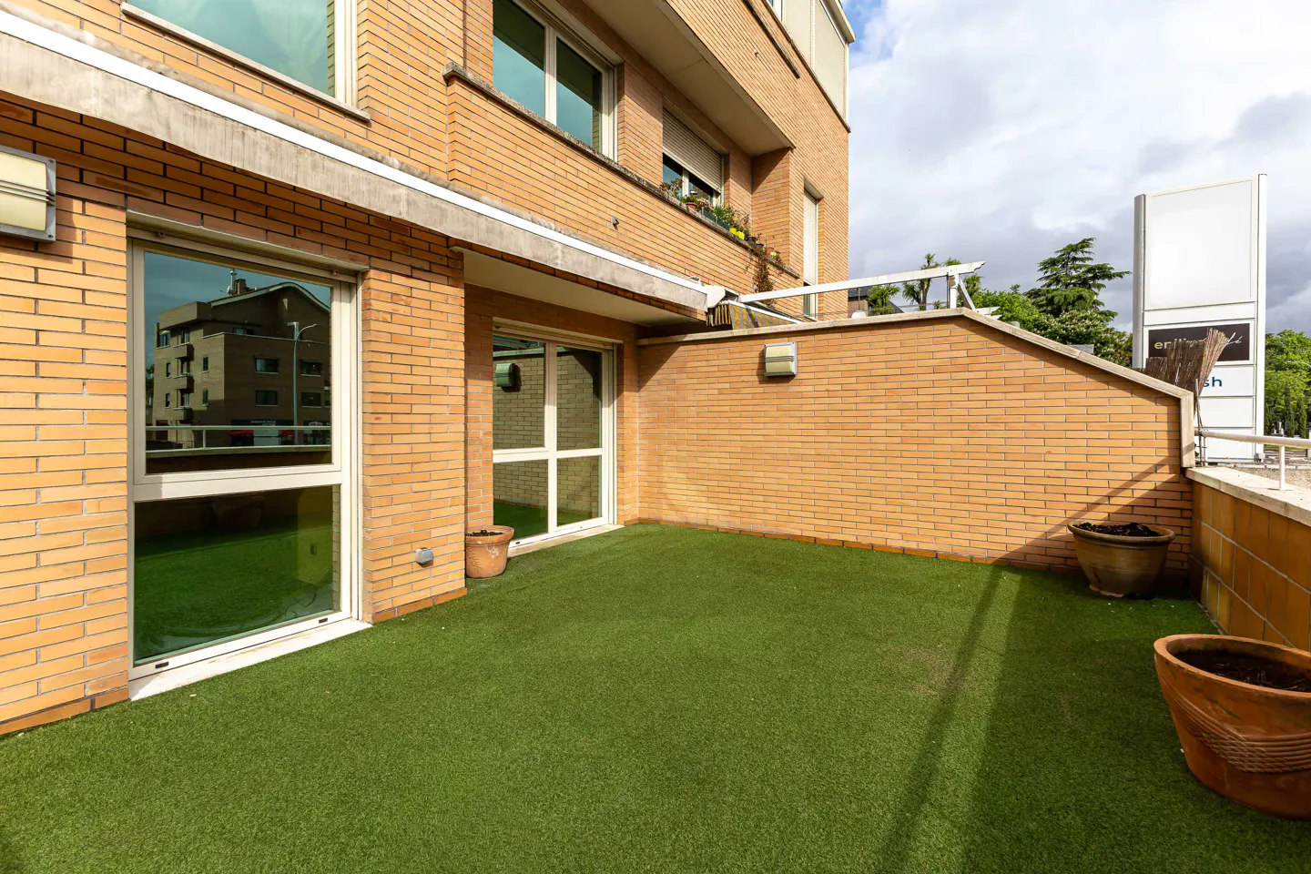 A brick building's outdoor patio with green turf, windows, and potted plants.