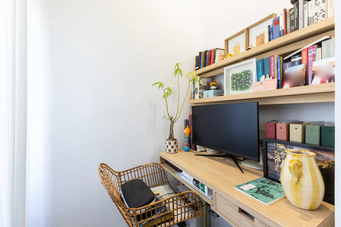 Bright home office with a wooden desk, black monitor, and wicker chair. Shelves above hold books and decor. A plant adds a touch of nature.