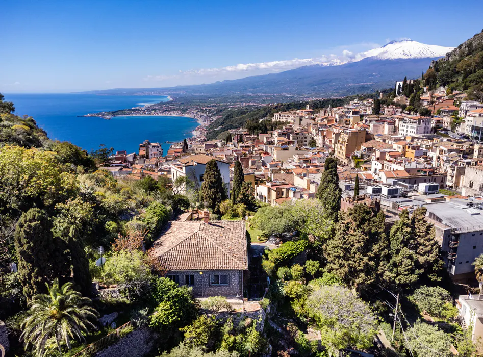 Scenic view of Taormina, Italy, with a stone house in the foreground, the Ionian Sea, and Mount Etna in the background.