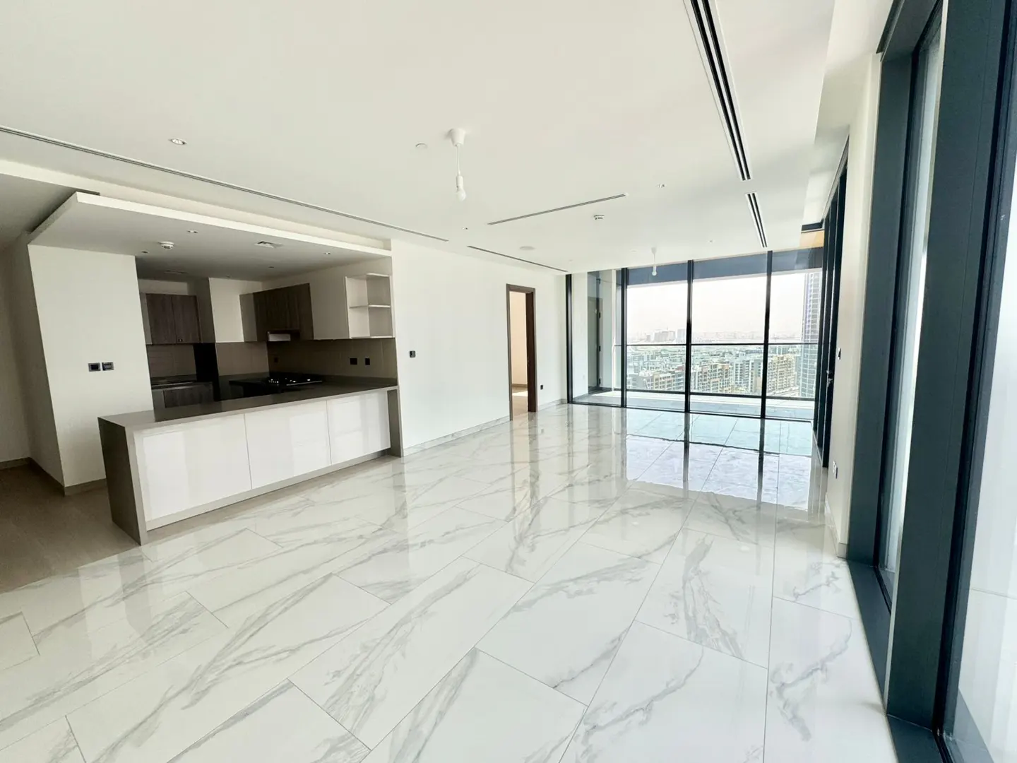 Bright, empty apartment with marble floors, floor-to-ceiling windows, and a modern kitchen with white cabinets. City view visible.