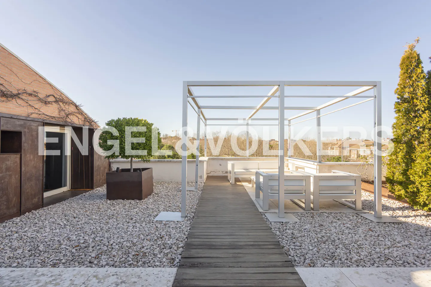 Rooftop terrace with a wooden walkway, white pergola, outdoor seating, gravel ground, and a small tree in a brown planter.