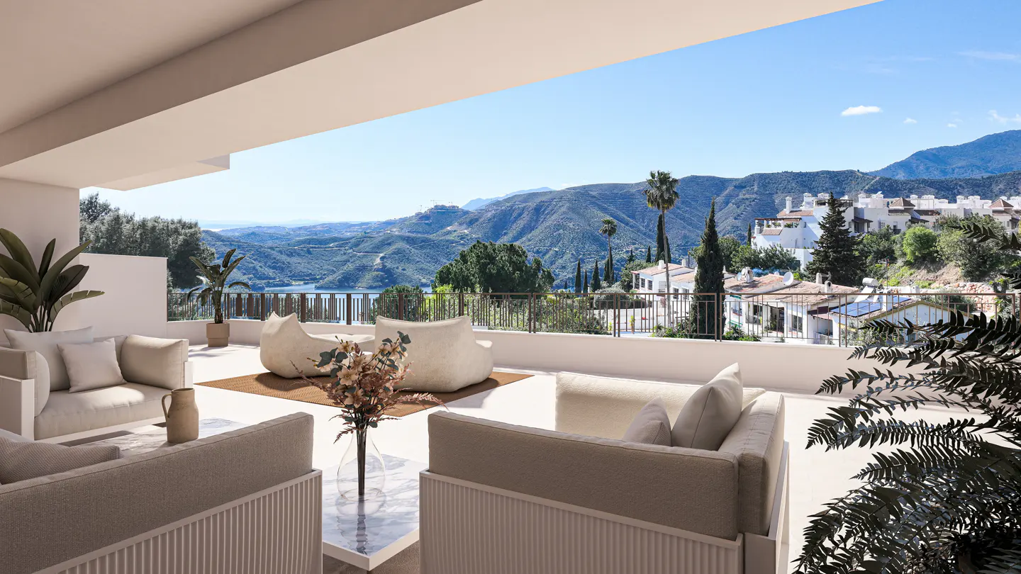 Outdoor patio with beige sofas, beanbag chairs, and a view of mountains, a lake, and white buildings under a blue sky.