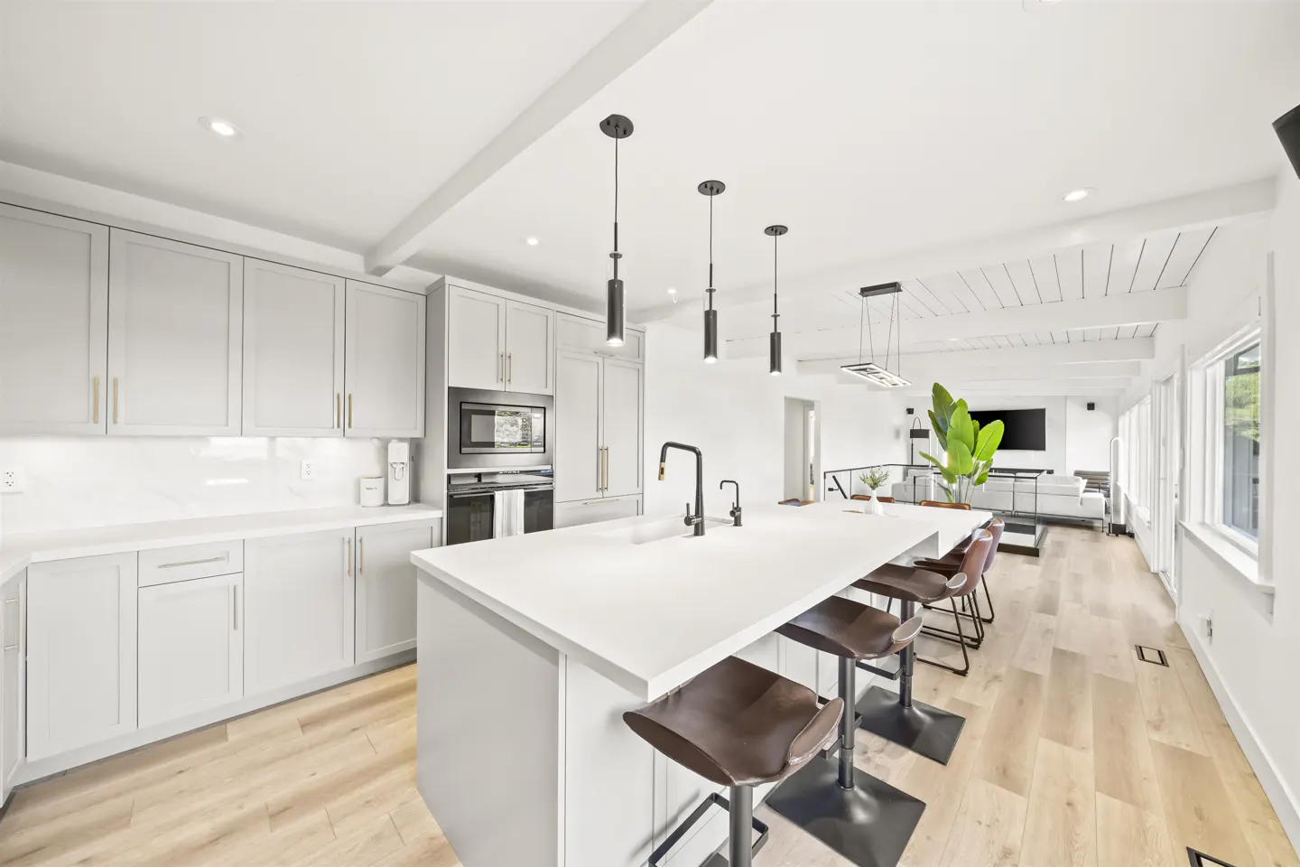 Bright, modern kitchen with gray cabinets, white countertops, and a large island with brown leather stools. Open floor plan leads to a living area.