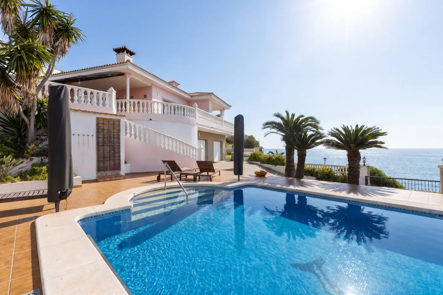 Luxury pink villa with a blue pool, palm trees, and ocean view on a sunny day. Two lounge chairs sit near the pool.