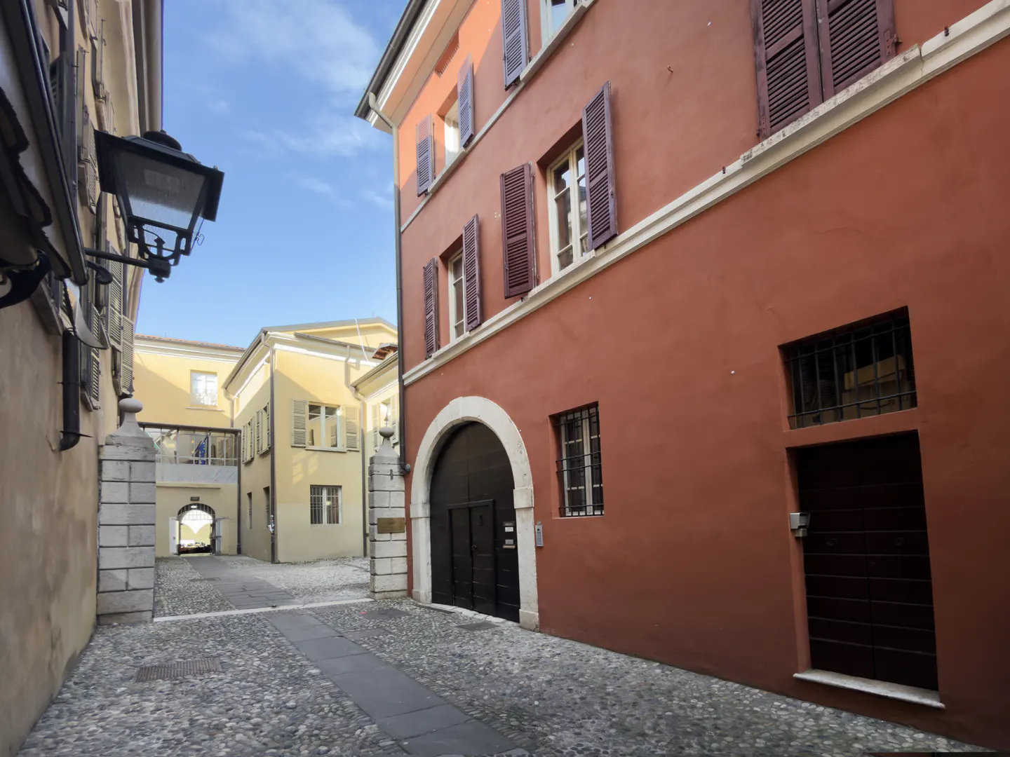 Cobblestone street view of a red building with brown shutters and a black arched doorway. A yellow building is visible in the background.