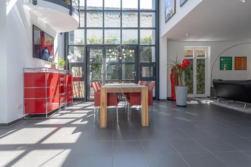 Bright, modern dining room with a wood table, red chairs, and a large window wall. Red cabinet and black sofa are also visible.