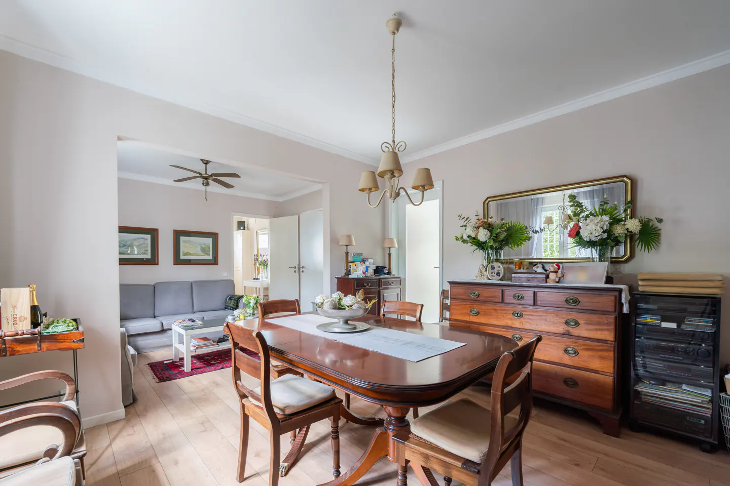 A dining room with a wooden table, chairs, and a chandelier. A living room is visible in the background.
