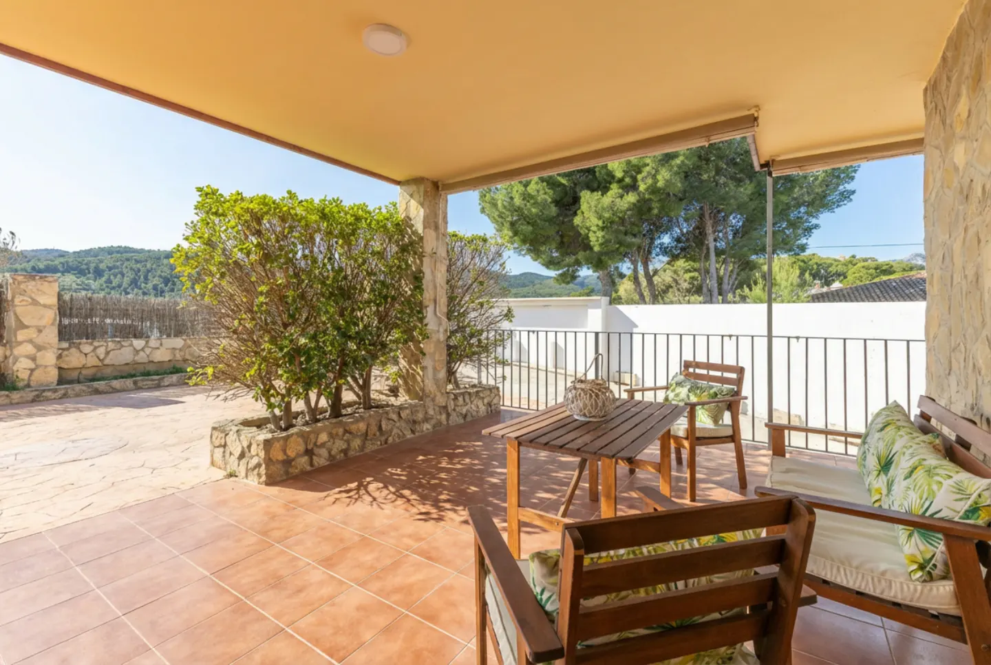 Covered patio with brown tile floor, stone walls, and a yellow ceiling. Wooden table and chairs with floral cushions sit near a black metal railing. Green trees and a blue sky are visible in the background.