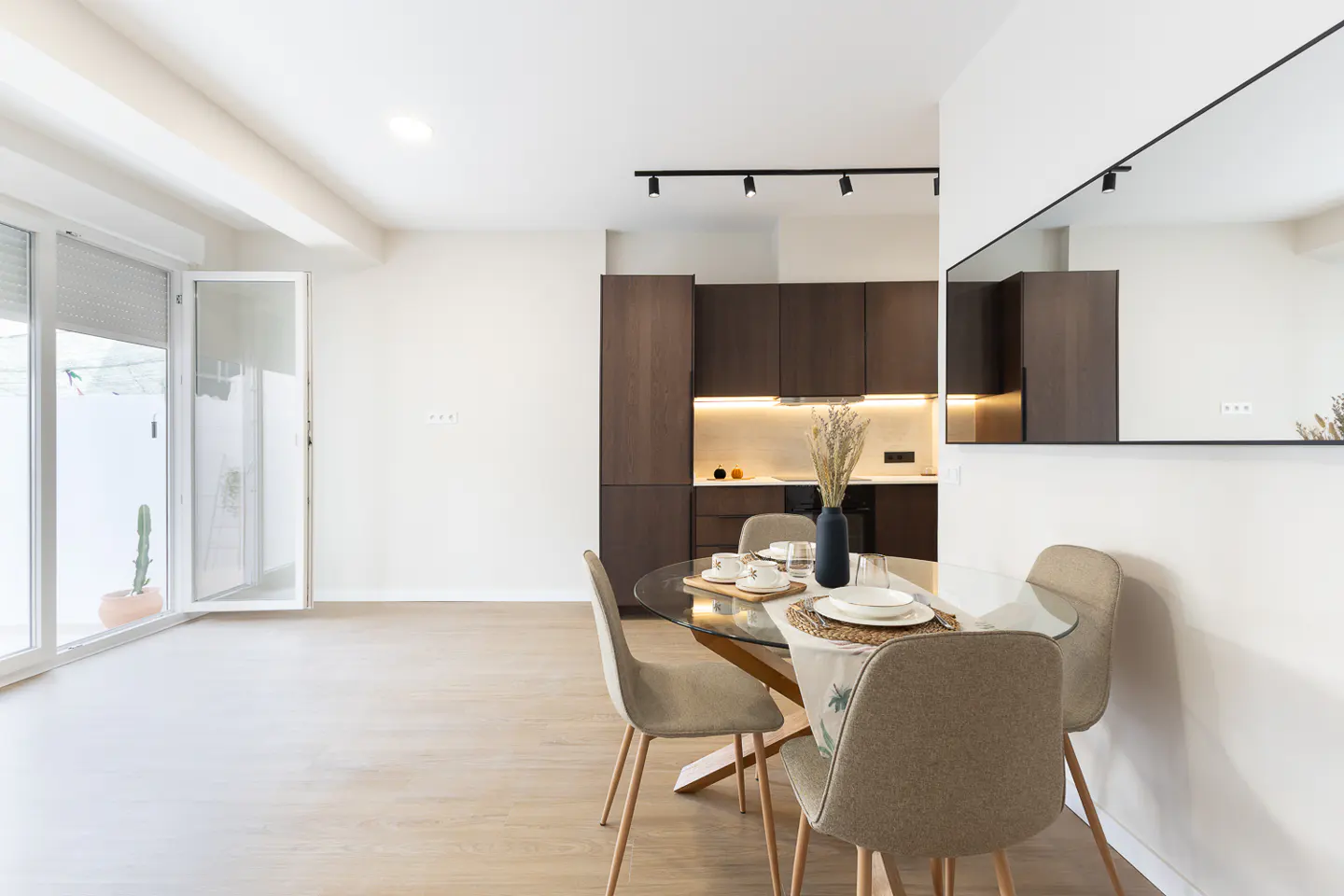 Bright apartment interior with a glass table set for dining, surrounded by four chairs. Dark wood cabinets line the kitchen area. A large window lets in natural light.