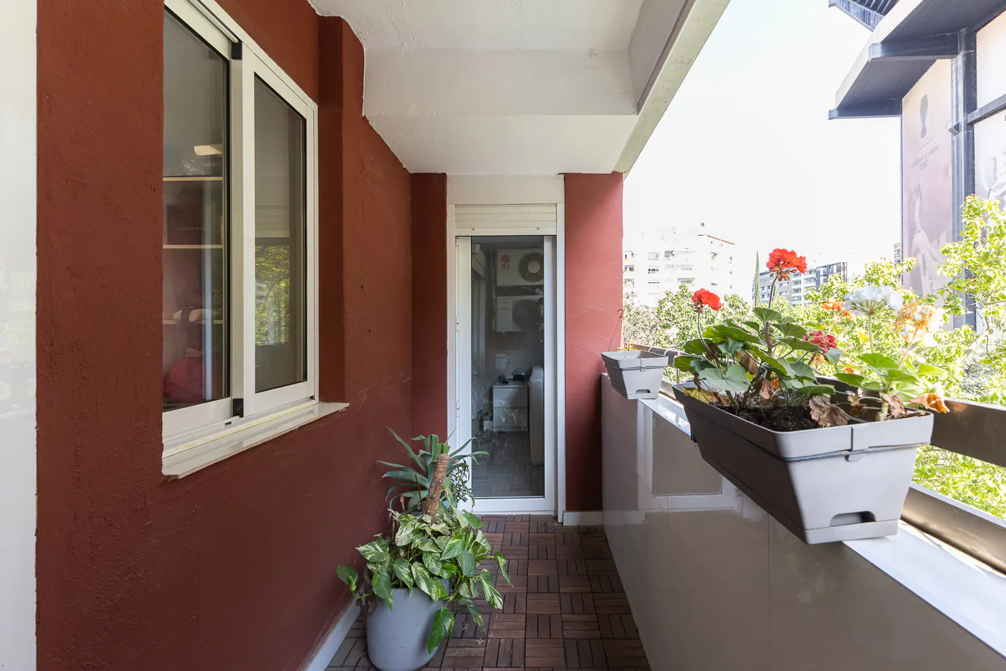 A balcony with red walls, a white ceiling, and a wood-tiled floor. Potted plants and flowers adorn the railing. A window and door lead inside.