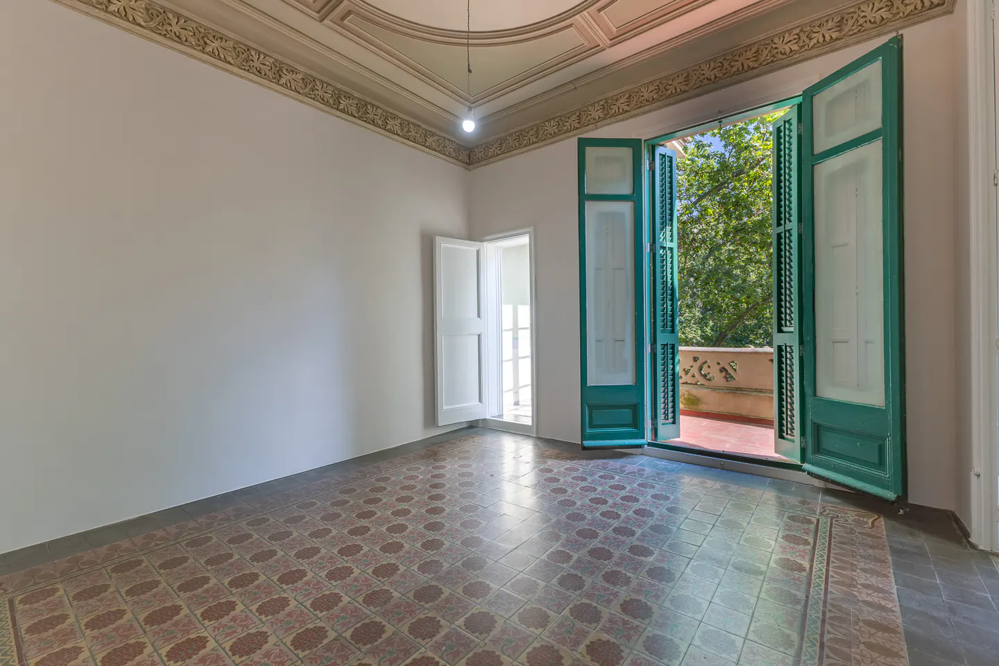 Empty room with patterned tile floor, white walls, and decorative ceiling. Open green shutters lead to a balcony with trees.