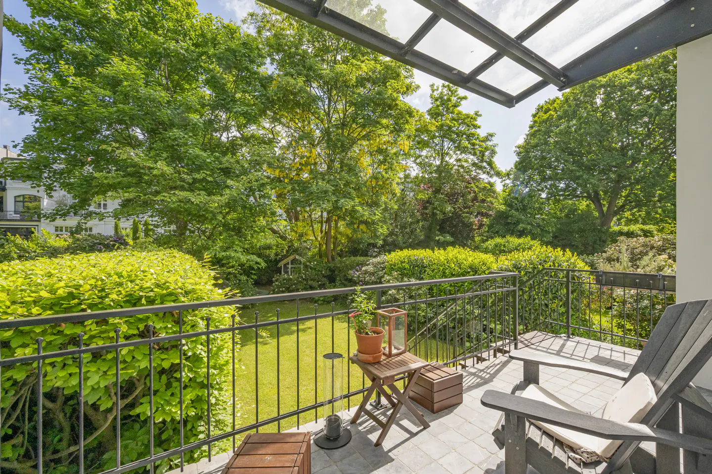 A balcony with a dark grey chair, a small wooden table with a potted plant, and a view of a lush green garden.