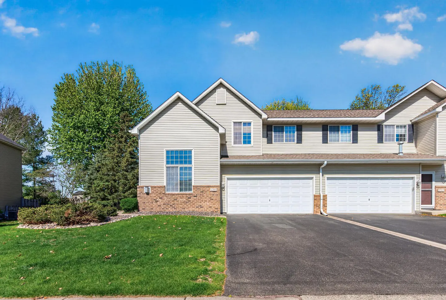 Beige two-story townhouse with two-car garage, asphalt driveway, green lawn, and blue sky.