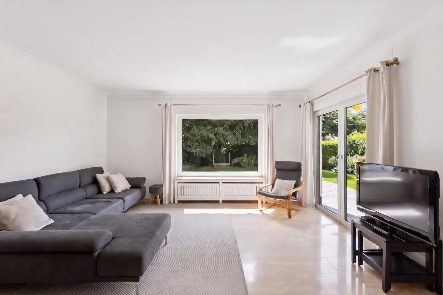 Bright living room with a grey sectional sofa, an armchair, a TV, and large windows overlooking a lush green garden.