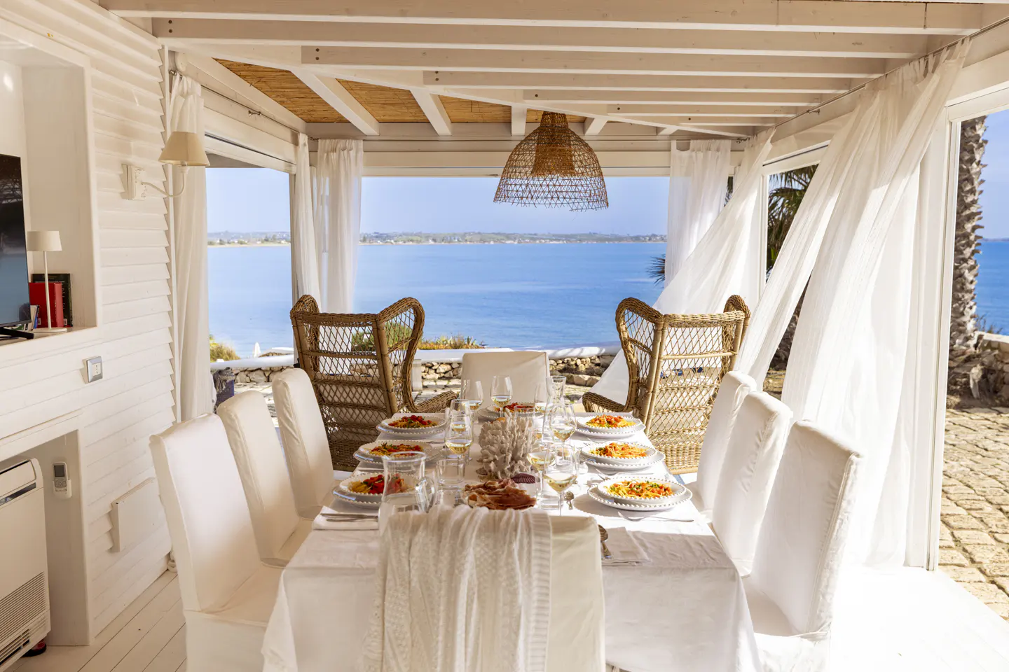 Outdoor dining area with a white table set for a meal, wicker chairs, and a view of the blue ocean.