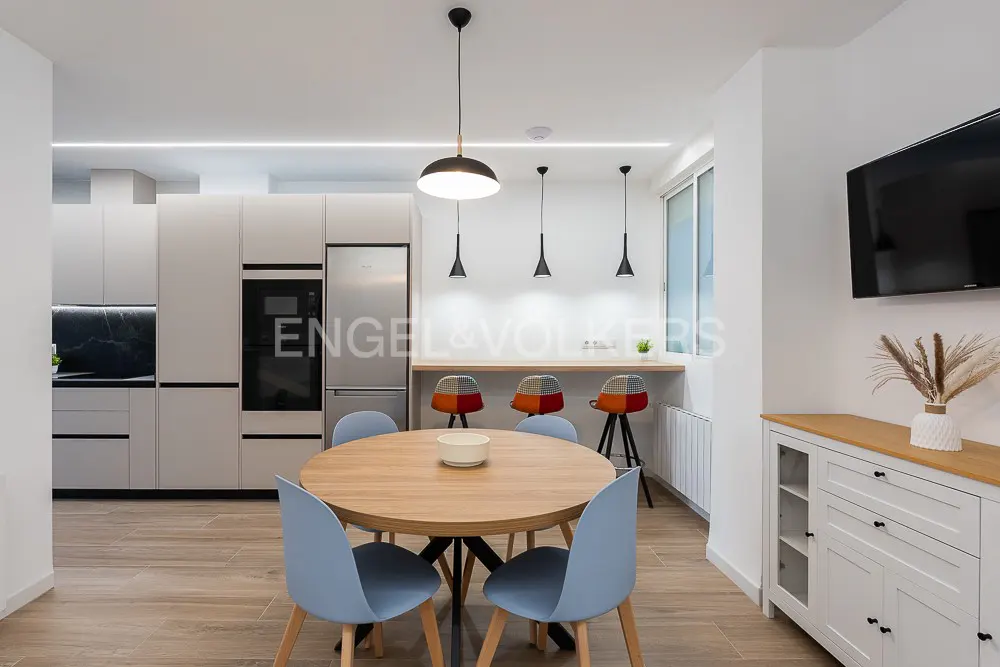 Bright, modern kitchen and dining area. A round wood table with blue chairs sits near a kitchen with gray cabinets and stainless steel appliances.