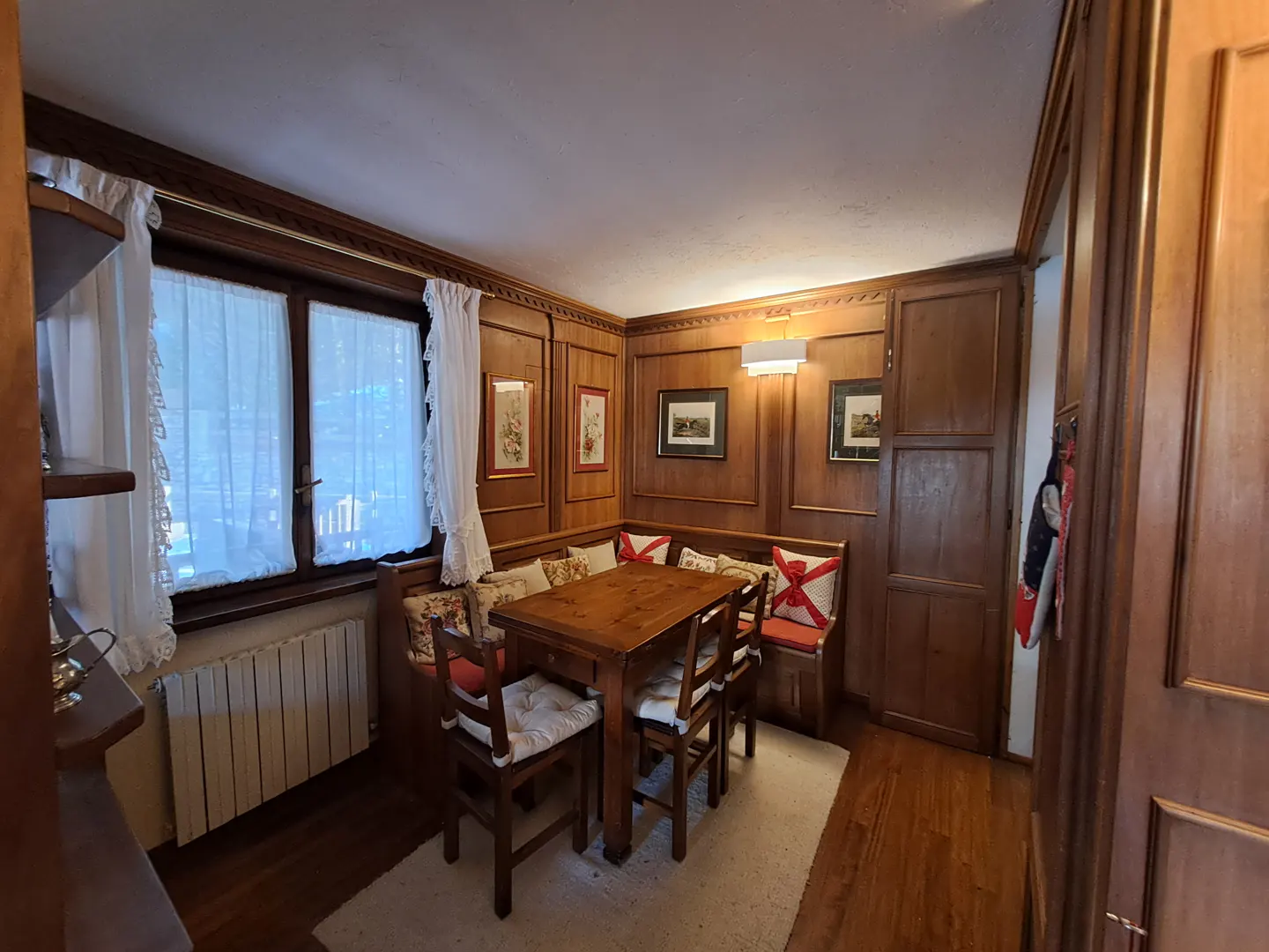 A cozy dining area with wood paneling, a table with chairs, and a cushioned corner bench. White curtains adorn the windows.