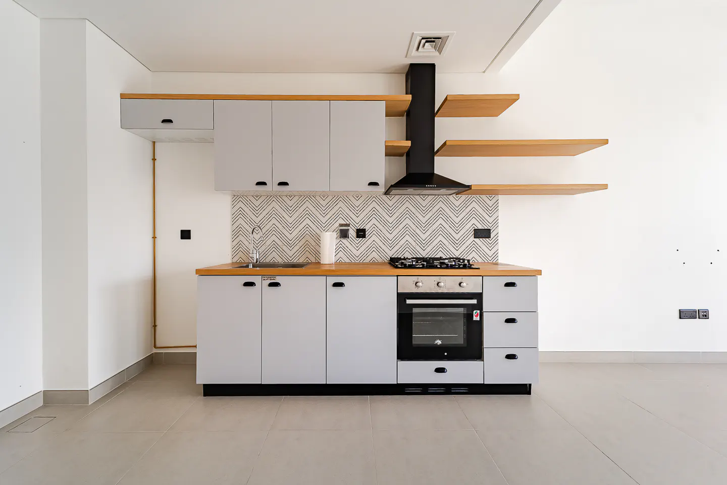 A modern kitchen with gray cabinets, wood countertops, and open shelves. A black range hood is above the stove. The backsplash has a chevron pattern.