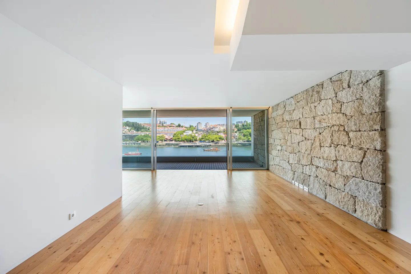 Bright, empty room with wood floors, white walls, and a stone accent wall. Sliding glass doors open to a balcony with a city and river view.