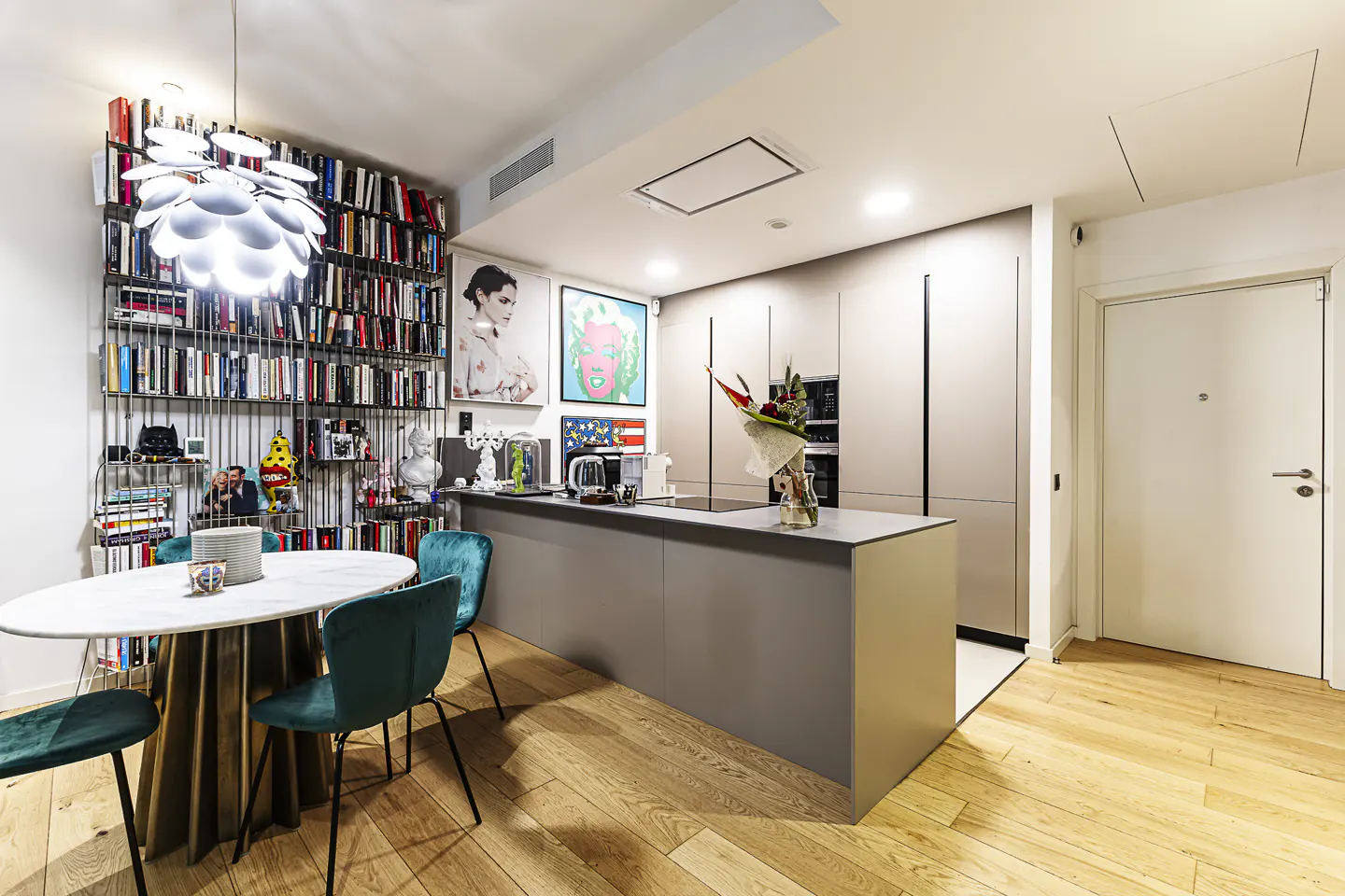 A modern kitchen with a bookshelf wall, a round table with teal chairs, and a gray kitchen island.