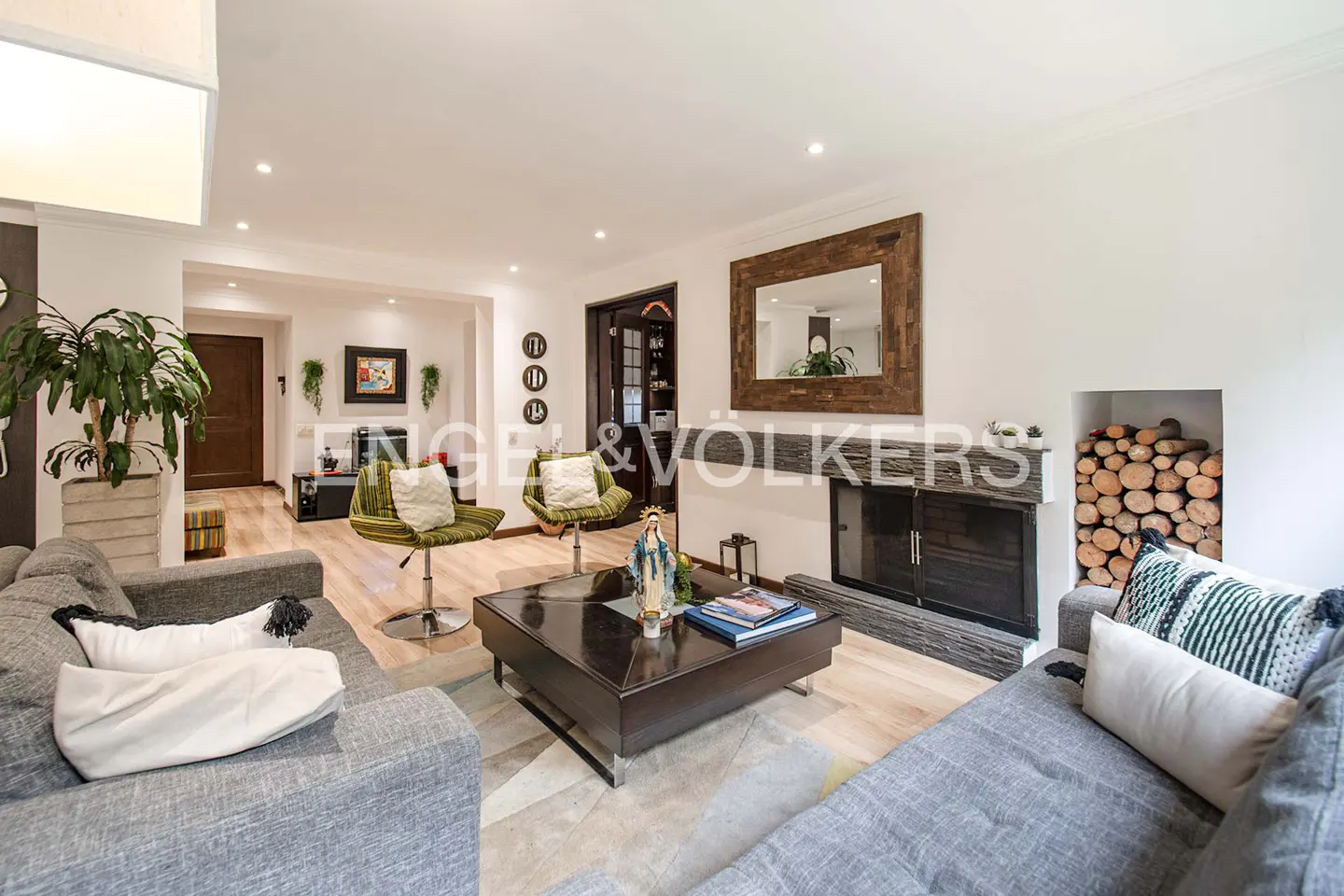 Living room with gray sofas, green chairs, and a dark wood fireplace with a mirror above it. A statue sits on the coffee table.