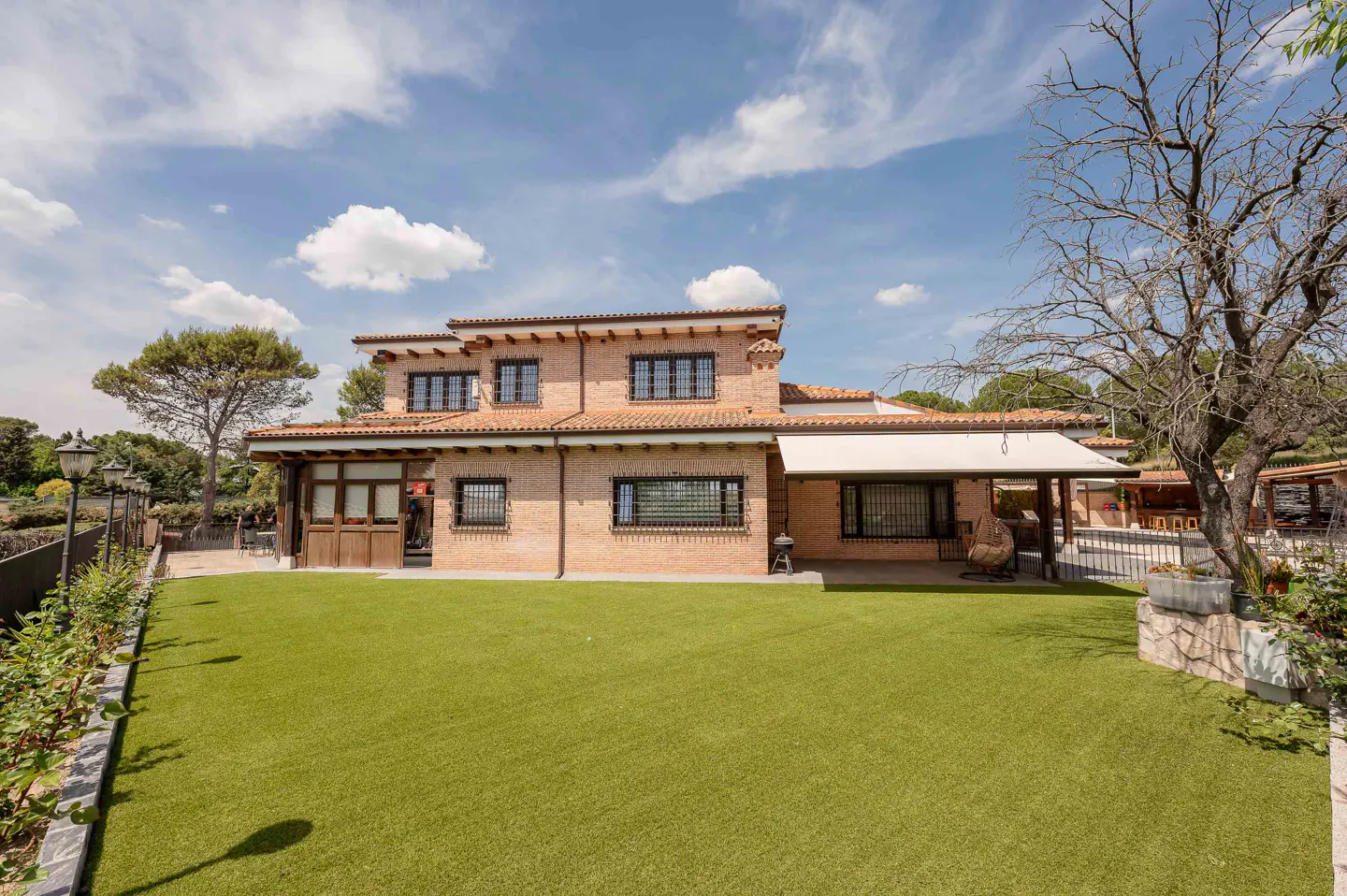 Two-story brick house with a red tile roof and black-framed windows, surrounded by a green lawn and trees under a blue sky.
