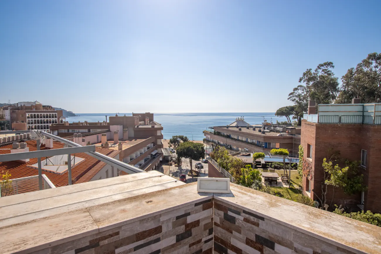 View from a balcony overlooking a coastal town with buildings, trees, and the ocean under a clear blue sky.