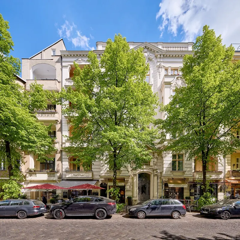 Elegant white building facade with ornate details, framed by green trees. Cars parked along the cobblestone street.
